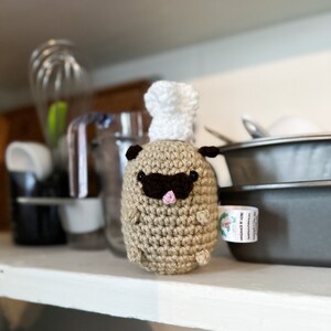 May include: A crocheted pug doll wearing a white chef's hat and a dark brown mustache. The pug has small black ears and a pink mouth. The doll is made of tan yarn and has a small tag attached.