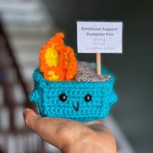 May include: A handmade crochet dumpster fire in teal, gray, orange, and yellow. The dumpster has a smiling face and a sign that reads "Emotional Support Dumpster Fire. It's fine. I'm fine. Everything is fine."