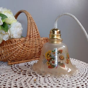 Può includere: Una lampada a sospensione in stile vintage con un motivo floreale. Il lampadario ha un bordo smerlato e una base color oro. Il paralume presenta un bouquet di fiori gialli, bianchi e arancioni. Un cestino di vimini e fiori bianchi sono sullo sfondo.