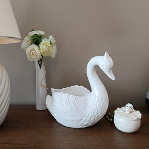 Può includere: Una fioriera a forma di cigno in ceramica bianca con dettagliate trame di piume, un lungo collo curvo e un'ampia apertura. Accanto c'è un piccolo contenitore bianco con una figura simile a una bambola sopra. È presente anche un vaso bianco con fiori.