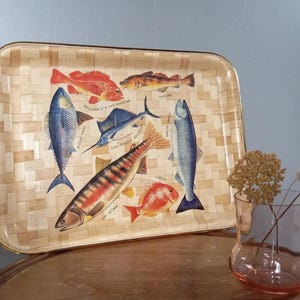 May include: Rectangular serving tray with a woven pattern and gold trim. The tray features illustrations of various fish species in shades of red, blue, and orange. A clear vase with dried flowers sits nearby.