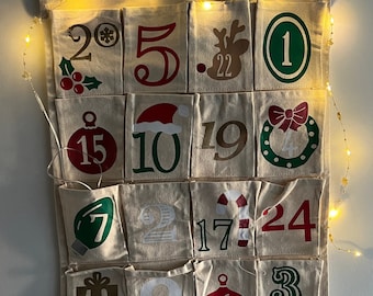 May include: A beige fabric advent calendar with 24 numbered pockets, each with a festive Christmas design. The top reads "Your Name Here". Fairy lights are strung along the top. A decorative piece for the holiday season.
