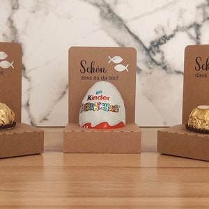 Könnte beinhalten: Drei braune Pappkartons mit einem kleinen Schokoladenei oder Bonbon in jedem. Die Kartons haben ein weißes Fischsymbol und den Text "Schon dass du da bist!" auf der Vorderseite.