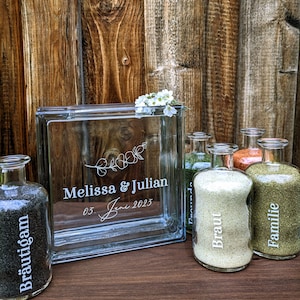 Puede incluir: Un bloque de vidrio con la inscripción "Melissa & Julian 03 Juni 2023" y un diseño floral. Cinco botellas de vidrio con arena están dispuestas frente al bloque. Las botellas están etiquetadas como "Braut", "Familie", "Brautigan" y "Trauzeugen".