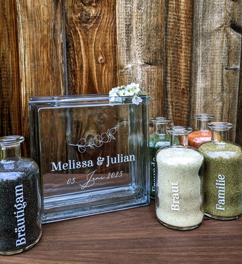 May include: A glass block with the inscription "Melissa & Julian 03 Juni 2023" and a floral design. Five glass bottles with sand are arranged in front of the block. The bottles are labeled "Braut", "Familie", "Brautigan", and "Trauzeugen".