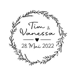 May include: A black and white illustration of a floral wreath with the text "Tim & Vanessa" and "28. Mai 2022" inside the wreath.