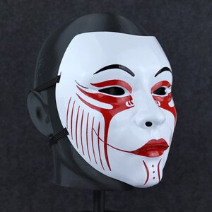 May include: White plastic mask with red and black details. The mask has a stylised face with a wide smile and red lines around the eyes and mouth.