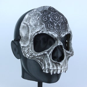 Pode incluir: Uma m&aacute;scara de caveira branca com um desenho de mandala preto na testa. A m&aacute;scara tem orif&iacute;cios para os olhos e uma abertura para a boca.