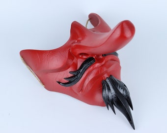 Pode incluir: Uma máscara japonesa Oni vermelha e preta com um nariz comprido e uma barba pontiaguda. A máscara é feita de um material texturizado e tem um design tradicional.