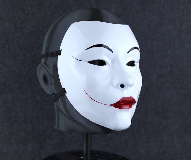 May include: A white mask with a black outline around the eyes and a red smile. The mask is on a black stand.