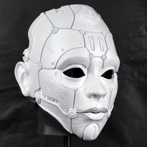Op de afbeelding: Een wit, futuristisch masker met een gedetailleerd, mechanisch ontwerp. Het masker heeft een glad gezicht met gedefinieerde kenmerken, waaronder een neus, lippen en oogkassen. Ingewikkelde lijnen en cirkelvormige accenten dragen bij aan de robotachtige esthetiek. Het masker wordt tegen een donkere achtergrond gepresenteerd.