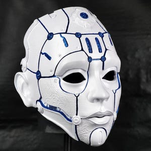 Op de afbeelding: Een wit masker met een robotachtig ontwerp, met blauwe accenten langs de randen en details. Het masker heeft een gladde, matte afwerking en is ontworpen met een menselijk gezicht, inclusief oogkassen, een neus en een mond.