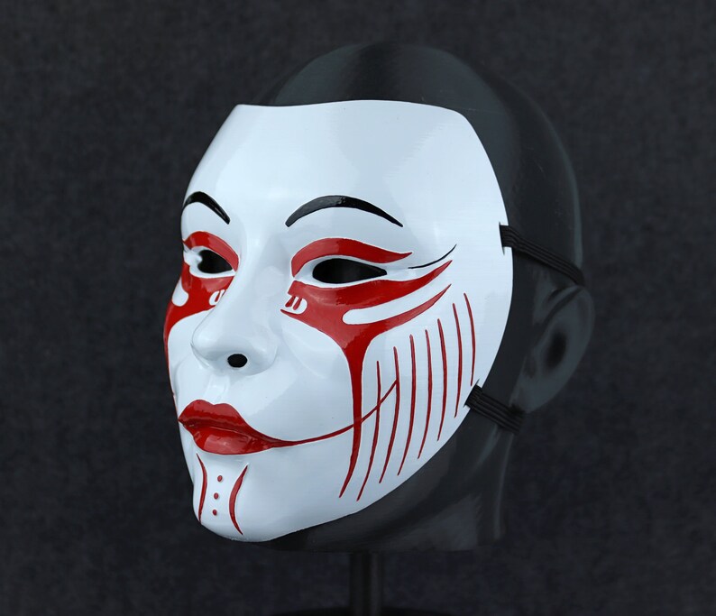 May include: A white mask with red and black details. The mask has a stylised face with a wide smile and a black eye line. The mask is on a black background.