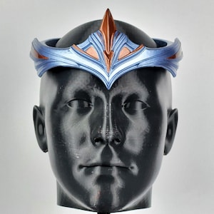 May include: A blue and copper-colored crown with a central pointed design, displayed on a black mannequin head. The crown features a metallic finish and intricate detailing, suitable for cosplay or costume events.