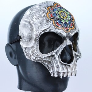 Pode incluir: Uma m&aacute;scara de caveira branca com um design de mandala colorido na testa. A m&aacute;scara tem orif&iacute;cios para os olhos e uma abertura para a boca.