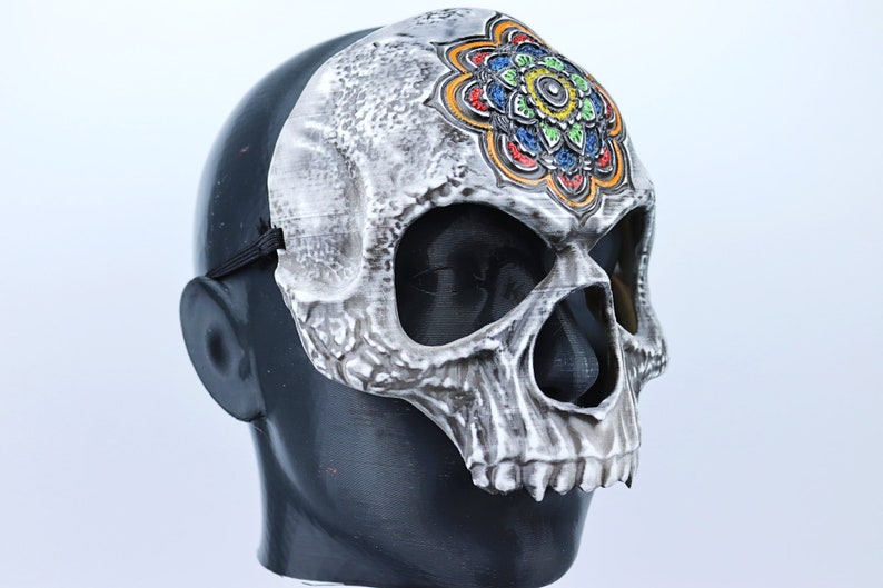 Pode incluir: Uma m&aacute;scara de caveira branca com um design de mandala colorido na testa. A m&aacute;scara tem orif&iacute;cios para os olhos e uma abertura para a boca.