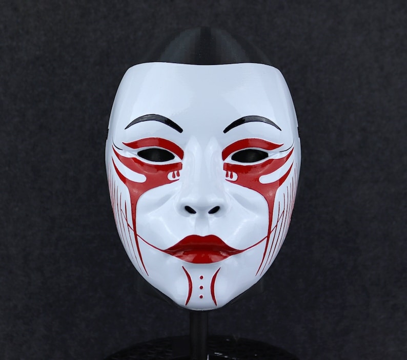 May include: White mask with red and black painted designs. The mask has a stylised human face with a black line for eyebrows and red lines for eyes and mouth.
