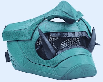 May include: A teal green and black plastic mask with a textured surface and a mesh mouth guard. The mask has a sharp, angular design with a pointed nose and two black horns.