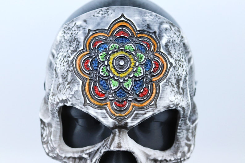 Pode incluir: Uma m&aacute;scara de caveira branca com um design de mandala colorido na testa. O mandala &eacute; pintado em tons de vermelho, laranja, amarelo, verde, azul e preto.