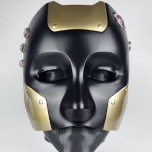 May include: A black mask with gold-colored accents. The mask has a smooth, reflective surface with gold plates on the forehead and cheeks. Small, pinkish-purple details are visible on the sides. The mask is designed to cover the face.