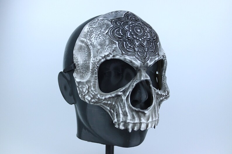 Pode incluir: Uma m&aacute;scara de caveira branca com um desenho de mandala preto na testa. A m&aacute;scara tem orif&iacute;cios para os olhos e uma abertura para a boca.