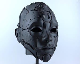 May include: A black, detailed, and intricate mask with a human-like face. The mask is made of a hard material and has a textured surface. It features a series of metal plates and bolts, giving it a futuristic and robotic look.
