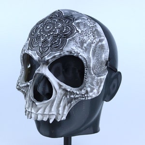 Pode incluir: Uma m&aacute;scara de caveira branca com um design de mandala preto na testa. A m&aacute;scara tem orif&iacute;cios para os olhos e uma abertura para a boca. A m&aacute;scara est&aacute; em uma cabe&ccedil;a de manequim preta.