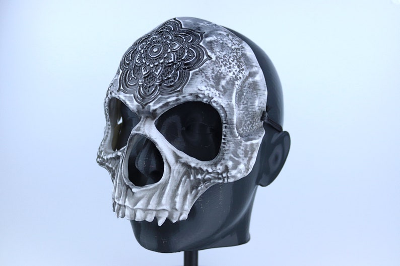 Pode incluir: Uma m&aacute;scara de caveira branca com um design de mandala preto na testa. A m&aacute;scara tem orif&iacute;cios para os olhos e uma abertura para a boca. A m&aacute;scara est&aacute; em uma cabe&ccedil;a de manequim preta.