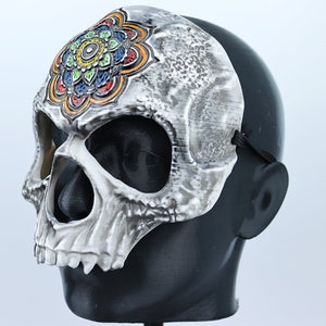 Pode incluir: Uma m&aacute;scara de caveira branca com um design de mandala colorido na testa. A m&aacute;scara tem tiras el&aacute;sticas pretas para usar.