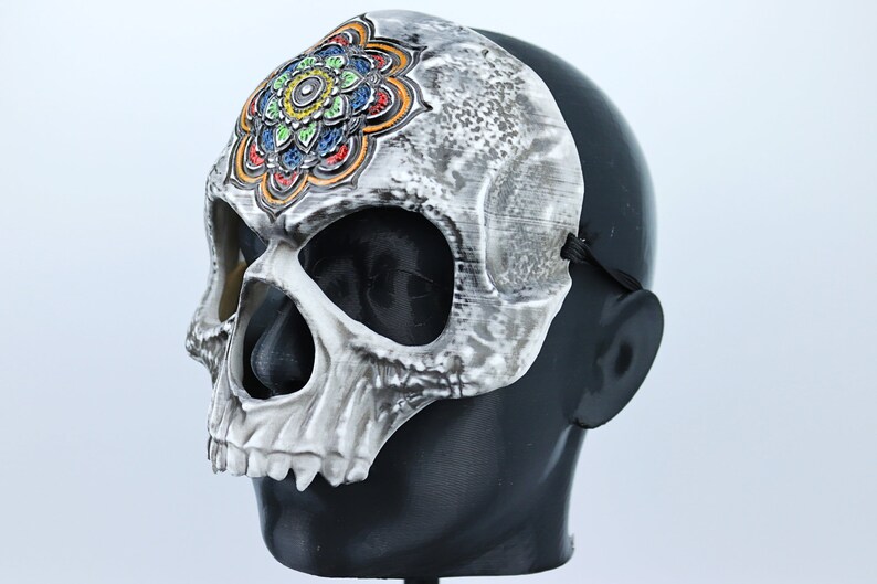 Pode incluir: Uma m&aacute;scara de caveira branca com um design de mandala colorido na testa. A m&aacute;scara tem tiras el&aacute;sticas pretas para usar.