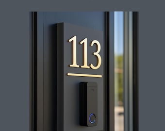 May include: A rectangular dark gray doorbell panel featuring the house number 113 in large gold-colored metallic numerals. Below the number is a horizontal gold line and a black rectangular doorbell device with a circular blue light indicator.