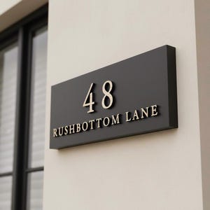 May include: A rectangular black house sign featuring the number 48 and the text RUSHBOTTOM LANE in raised gold-toned lettering. The sign is mounted horizontally on a light-coloured wall, displaying a clean, modern, and high-contrast design.