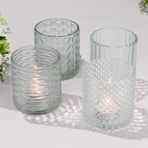 Könnte beinhalten: Vier klare Glas-Teelichthalter mit unterschiedlichen Texturen. Einer hat ein wellenförmiges Muster, ein anderer ein Blattdesign, ein dritter ein gepunktetes Muster und der letzte vertikale Linien. Jeder ist zylindrisch und für eine kleine Kerze konzipiert.