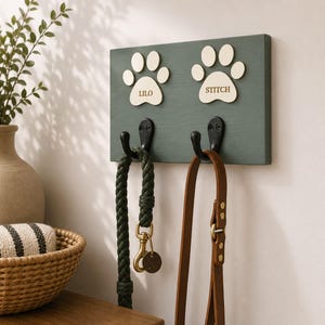 May include: A rectangular dark green wooden wall mount featuring two light-coloured paw print cutouts labelled LILO and STITCH. Two black metal hooks are attached to the board, holding a braided dark green lead and a brown leather lead.
