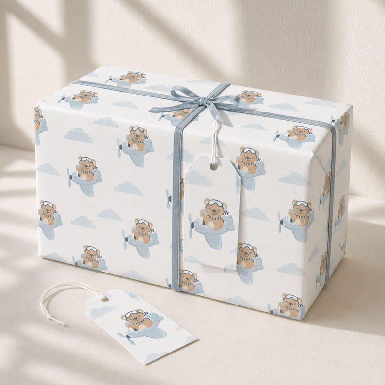 May include: A rectangular gift box wrapped in white paper featuring a repeating pattern of brown teddy bears wearing aviator goggles and flying light blue airplanes among light blue clouds. A light blue ribbon is tied around the box with a matching tag.