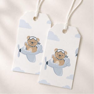 May include: Two rectangular white gift tags with rounded top corners and white string loops. Each tag features a brown teddy bear wearing aviator goggles and a striped scarf, flying a light blue airplane amidst light blue clouds.