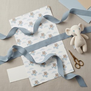 May include: A sheet of white wrapping paper featuring a repeating pattern of brown teddy bears flying light blue airplanes among light blue clouds, accompanied by a cream-colored knit elephant toy, gold scissors, and a long dusty blue ribbon.