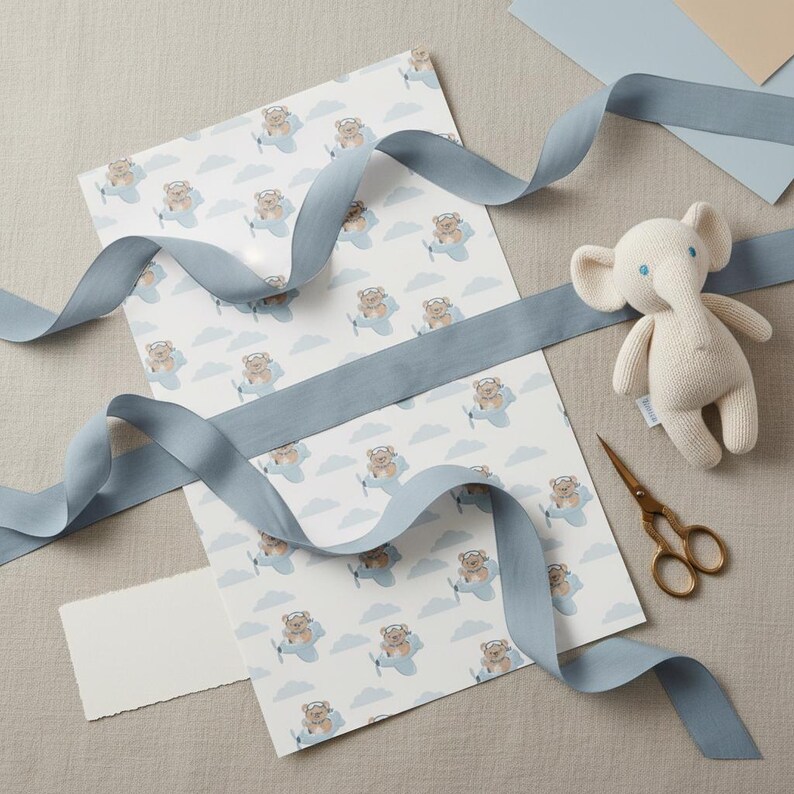 May include: A sheet of white wrapping paper featuring a repeating pattern of brown teddy bears flying light blue airplanes among light blue clouds, accompanied by a cream-colored knit elephant toy, gold scissors, and a long dusty blue ribbon.