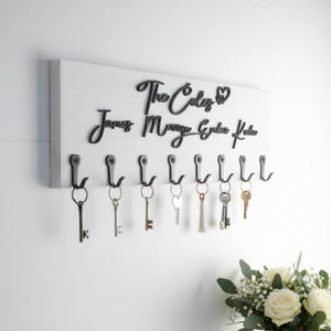 May include: A rectangular white wooden wall-mounted key holder featuring black script lettering that reads The Coleys followed by the names James, Morgan, Evelyn, and Kaden. It includes eight black metal hooks, with several keys hanging from them.