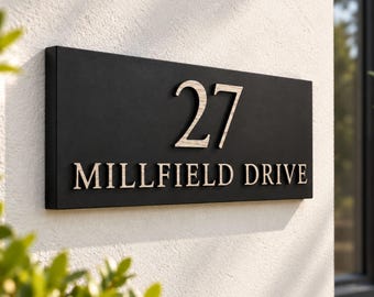 May include: A rectangular black house sign featuring the number 27 and the text MILLFIELD DRIVE in light-colored, raised lettering. The sign is mounted on a textured white wall, with green foliage visible in the foreground.