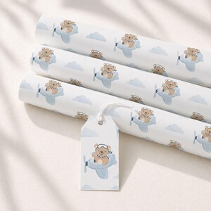 May include: Three rolls of white wrapping paper featuring a repeating pattern of brown teddy bears wearing aviator goggles and scarves while flying light blue airplanes among light blue clouds, accompanied by a matching rectangular gift tag.