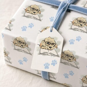 May include: A white gift box wrapped in paper featuring a repeating illustration of a light brown dog wearing black glasses and reading an open book, accented by light blue paw prints and tied with a light blue velvet ribbon and a matching gift tag.