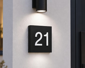 May include: A square black house number plaque featuring the white digits 21 mounted on a white wall. Above the plaque, a cylindrical black wall light fixture casts a downward glow, illuminating the surface of the sign and the wall below it.