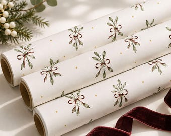 May include: Three rolls of off-white wrapping paper featuring a repeating pattern of green mistletoe sprigs tied with dark red bows and small gold dots. A length of deep burgundy velvet ribbon is positioned in the foreground of the paper rolls.