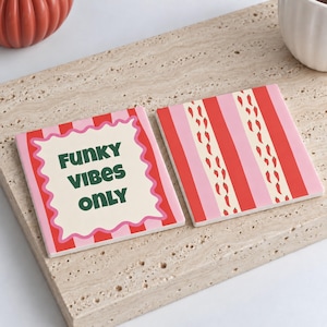May include: Two square coasters with a red and pink striped pattern. One coaster features a cream-colored wavy-edged center with the dark green text "FUNKY VIBES ONLY". The other coaster displays a repeating pattern of small red chili peppers.