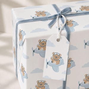 May include: A rectangular gift box wrapped in white paper featuring a repeating pattern of brown teddy bears wearing aviator goggles and scarves while flying light blue airplanes among light blue clouds, tied with a blue velvet ribbon and tag.