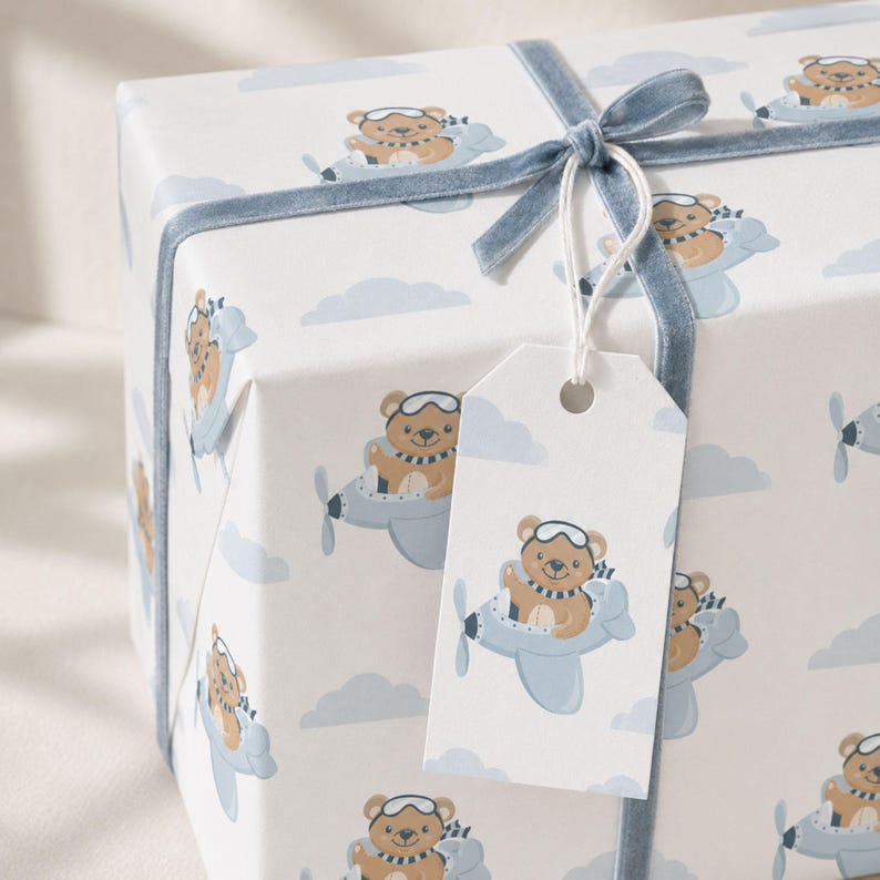 May include: A rectangular gift box wrapped in white paper featuring a repeating pattern of brown teddy bears wearing aviator goggles and scarves while flying light blue airplanes among light blue clouds, tied with a blue velvet ribbon and tag.