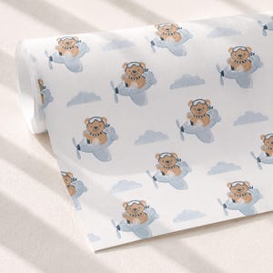 May include: A roll of white wrapping paper featuring a repeating pattern of brown teddy bears wearing aviator goggles and striped scarves, each piloting a light blue airplane amidst scattered light blue clouds on a clean white background.