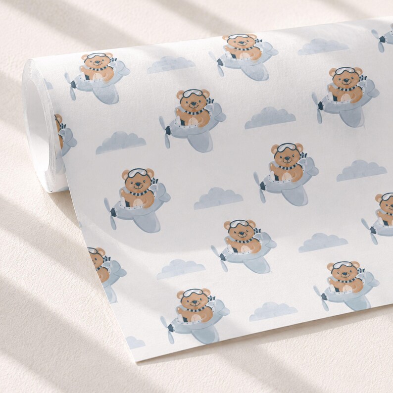 May include: A roll of white wrapping paper featuring a repeating pattern of brown teddy bears wearing aviator goggles and striped scarves, each piloting a light blue airplane amidst scattered light blue clouds on a clean white background.