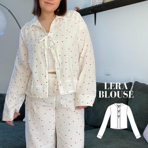 Könnte beinhalten: Eine weiße Bluse mit rotem Herzprint und einer Schleife am Hals. Die Bluse ist Teil eines passenden Sets mit einer Hose. Das Bild zeigt auch eine Strichzeichnung der Bluse mit dem Text "Lera Blouse".
